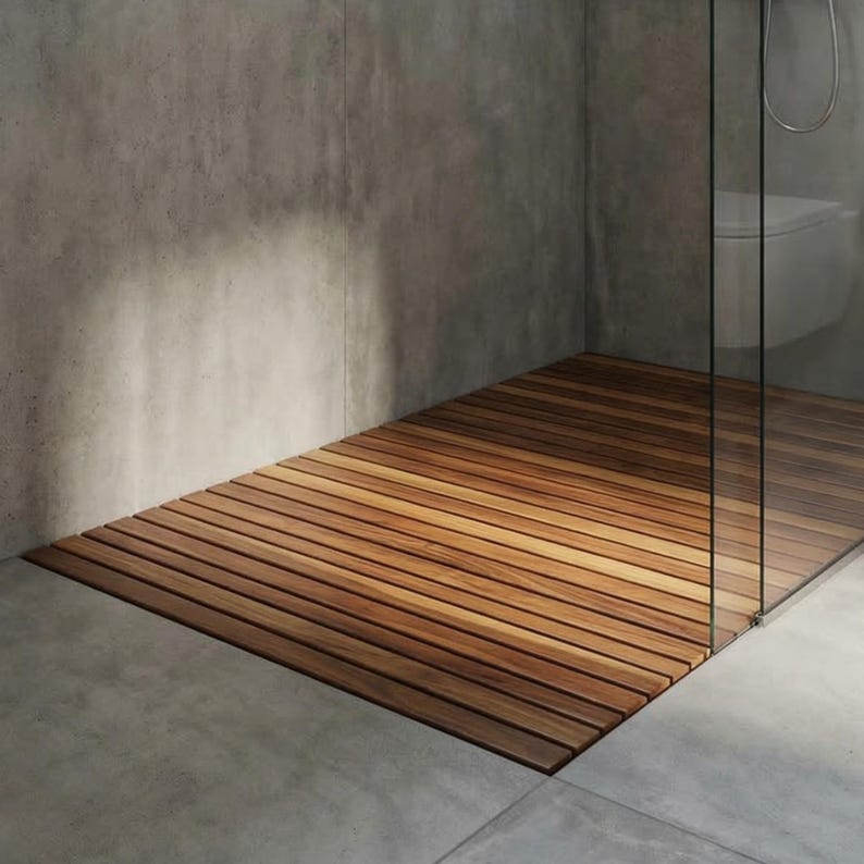 May include: A wooden shower mat made of parallel wooden slats. The mat is a warm brown color and sits on a gray concrete floor. The shower stall has a glass door and a gray concrete wall.
