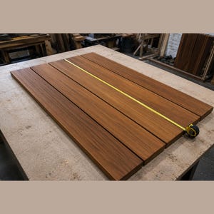 May include: Four rectangular wooden planks with a smooth, medium-brown grain finish arranged side-by-side. A yellow retractable measuring tape is extended diagonally across the surface of the planks to show their length.