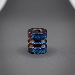 May include: A cylindrical metal bead featuring a threaded center hole and a grooved exterior. The surface displays an iridescent finish with swirling patterns of deep blue, purple, and bronze tones, creating a metallic, multi-colored appearance.