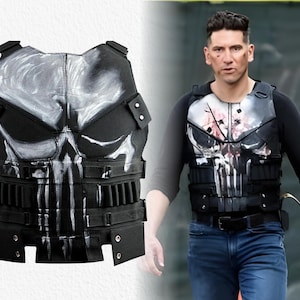 May include: Black tactical vest with a white skull design. The vest has multiple pouches and straps. It is worn over a black long-sleeve top. This is a costume piece.