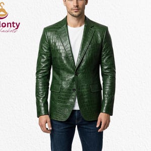 May include: A green leather blazer with a textured crocodile pattern. The blazer has a notched lapel, two front pockets, and a single-button closure. The model is wearing a white t-shirt and blue jeans. The logo "Monty Jackets" is in the top left corner.