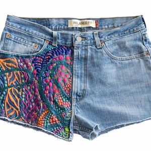 May include: Light blue denim shorts with a relaxed fit. The left side features intricate, colorful embroidery in shades of green, orange, pink, and blue. The shorts have a classic five-pocket design and a button closure.