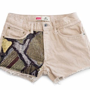 May include: Beige denim shorts with a frayed hem. The left front pocket features a patchwork design with a mix of patterns in black, green, and gold. The shorts have a button closure and classic five-pocket styling.