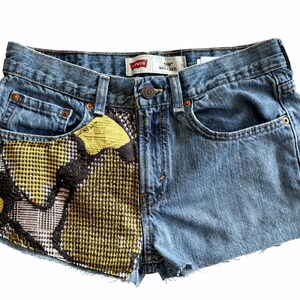 May include: Blue denim shorts with a unique design. The left pocket features a patchwork of yellow, black, and white patterned fabric. The shorts have a classic five-pocket design and a button closure. The Levi's brand label is visible.