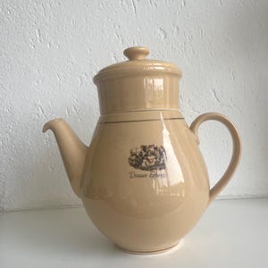 Op de afbeelding: Beige keramische theepot met deksel, tuit en handvat. De theepot heeft een afgeronde body en een decoratieve band aan de bovenkant. De woorden "Douwe Egberts" staan op de zijkant gedrukt, samen met een kleine illustratie.