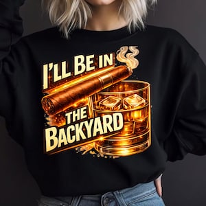 May include: Black sweatshirt with a graphic of a cigar, a glass of whiskey with ice cubes, and the text "I'll be in the backyard." The design is in shades of brown and gold.