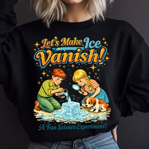 Pode incluir: Camisola preta com um gráfico colorido de duas crianças conduzindo uma experiência científica com gelo. O texto diz "Let's Make Ice Vanish!" e "A Fun Science Experiment!". Um cão pequeno também está presente.