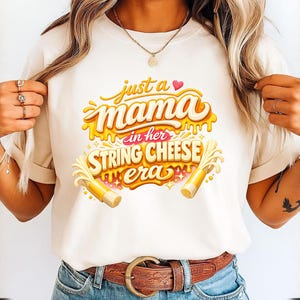 Peut inclure: T-shirt crème avec l'inscription "just a mama in her string cheese era" en jaune et orange. Le motif comprend un cœur et des illustrations de fromage en ficelle. Le t-shirt est porté avec un jean bleu et une ceinture marron.