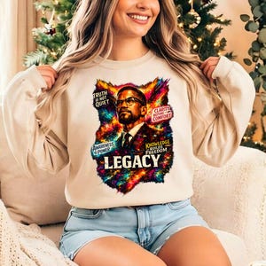 May include: A beige sweatshirt featuring a colorful graphic of a man with glasses, a tie, and the word "LEGACY". The graphic includes text that reads "TRUTH IS NOT QUIET", "AWARENESS IS POWER", "CLARITY BEFORE COMFORT", and "KNOWLEDGE BUILDS FREEDOM".