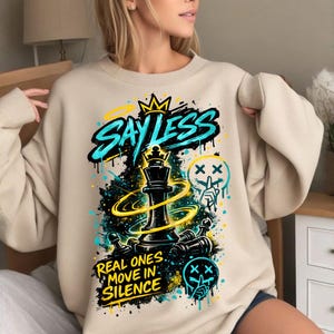 Puede incluir: Sudadera beige con un diseño gráfico que presenta una pieza de ajedrez de rey negro, las palabras "SAY LESS" en turquesa y amarillo, y la frase "REAL ONES MOVE IN SILENCE". El diseño incluye elementos de estilo grafiti.