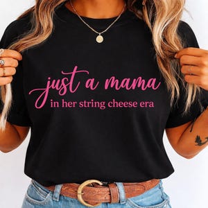 May include: Black t-shirt with the text "just a mama in her string cheese era" in pink script. The shirt is paired with blue jeans and a brown belt. A gold necklace and rings are visible.