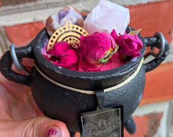 Puede incluir: Un pequeño caldero de hierro fundido negro lleno de cera rosa, capullos de rosa secos de color rosa intenso, un cristal morado claro, un cristal rosa pálido y un dije de metal dorado. Un colgante rectangular negro con un estampado gráfico blanco detallado cuelga del borde.