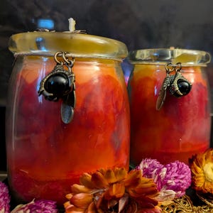 May include: Two jar candles with a red and orange gradient, topped with a clear lid. Each candle has a decorative charm with a black stone and a claw design. The candles are surrounded by dried flowers.