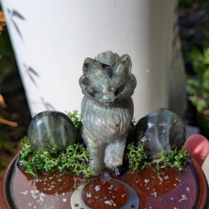 May include: A carved gray fox figurine with two matching spheres, set on a wooden base with green moss and a silver crescent moon design. The fox is detailed with visible fur and stands upright.