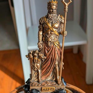 May include: Bronze-colored statue of Hades, the Greek god of the underworld, holding a trident and wearing a crown. The statue features a detailed depiction of Hades with a beard, and is accompanied by a three-headed dog, Cerberus. The base of the statue reads "HADES".