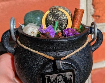Puede incluir: Un caldero de hierro fundido negro lleno de una piedra verde, un dije de media luna dorada, una rama de canela y flores secas moradas y blancas. Un colgante rectangular negro con la carta del tarot de la Rueda de la Fortuna cuelga de un cordel.