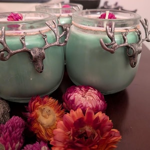 May include: Three teal-colored candles in glass jars, each adorned with a silver-toned deer antler design. The candles are decorated with dried flowers and sit on a dark surface. The candles have a rustic, nature-inspired theme.