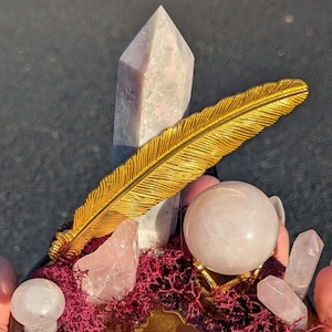 May include: A decorative display featuring a gold feather, rose quartz crystals, and a crystal ball on a wooden base with red moss. The arrangement includes a large quartz point and several smaller crystals.