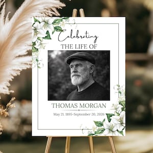 May include: A memorial sign on a wooden easel. The sign features a black and white photo of a man, surrounded by white lilies and green foliage. Text reads "Celebrating the Life of Thomas Morgan" with dates.