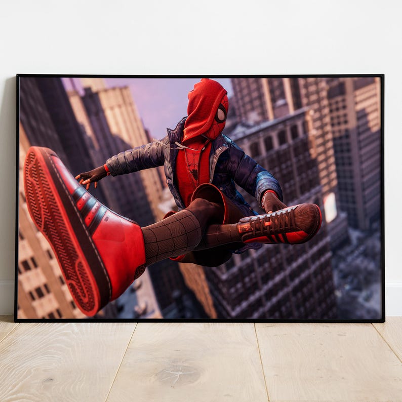 May include: Framed print of a superhero in a red and black suit, leaping through a city. The character wears a red hooded mask, a blue jacket, and red sneakers with black stripes. The background features tall buildings and a sunset.