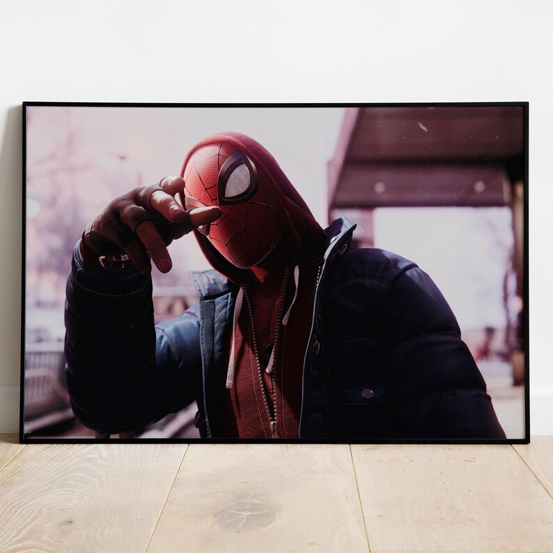 May include: Framed print featuring a person in a Spider-Man mask and red hoodie, wearing a black puffer jacket. The person is outdoors, with a blurred background of a building and trees. The image has a cinematic feel.