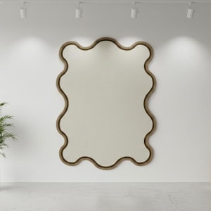 May include: A large, wavy-edged mirror with a light beige interior and a dark brown frame hangs on a white wall. A potted plant and a decorative vase on a wooden stand flank the mirror. The room is lit by overhead spotlights.