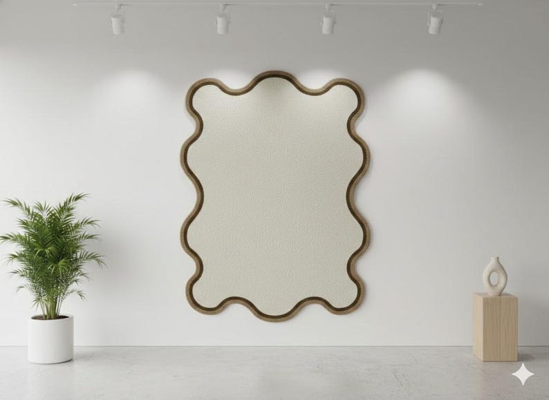 May include: A large, wavy-edged mirror with a light beige interior and a dark brown frame hangs on a white wall. A potted plant and a decorative vase on a wooden stand flank the mirror. The room is lit by overhead spotlights.