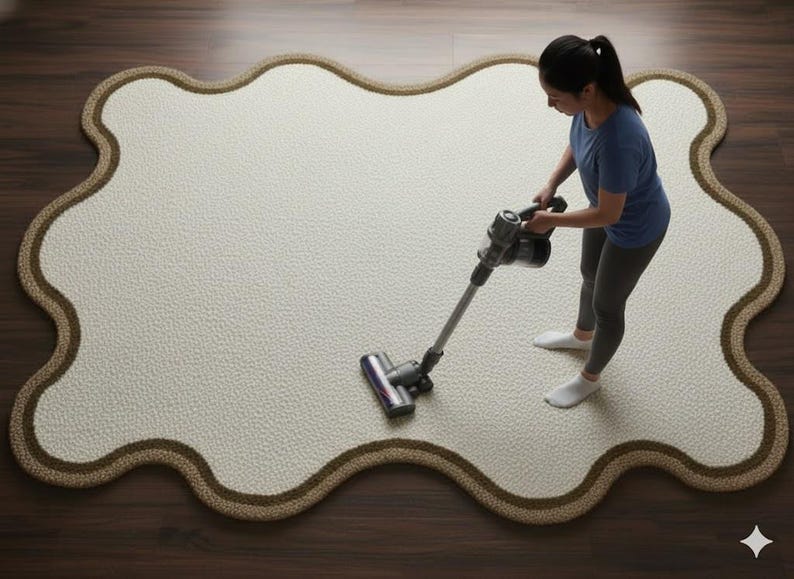 May include: A cream-colored, irregularly shaped rug with a brown border is being vacuumed by a person. The vacuum is gray and black. The rug is on a dark brown wooden floor.