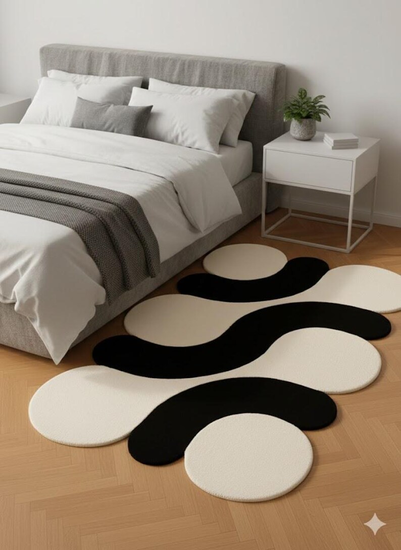 May include: A modern bedroom scene featuring a black and white abstract rug. The rug has a unique, flowing design with circular and wave-like shapes. The room includes a bed with white bedding and a gray headboard, and a white nightstand.