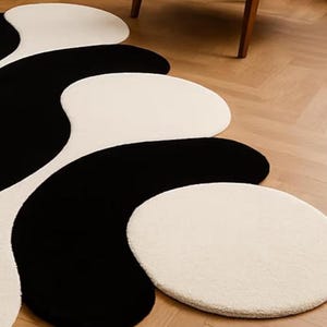 May include: A modern area rug with a unique design. The rug features a flowing pattern of black and off-white shapes. The shapes are soft and organic, creating a visually interesting and stylish floor covering. The rug is placed on a wooden floor.