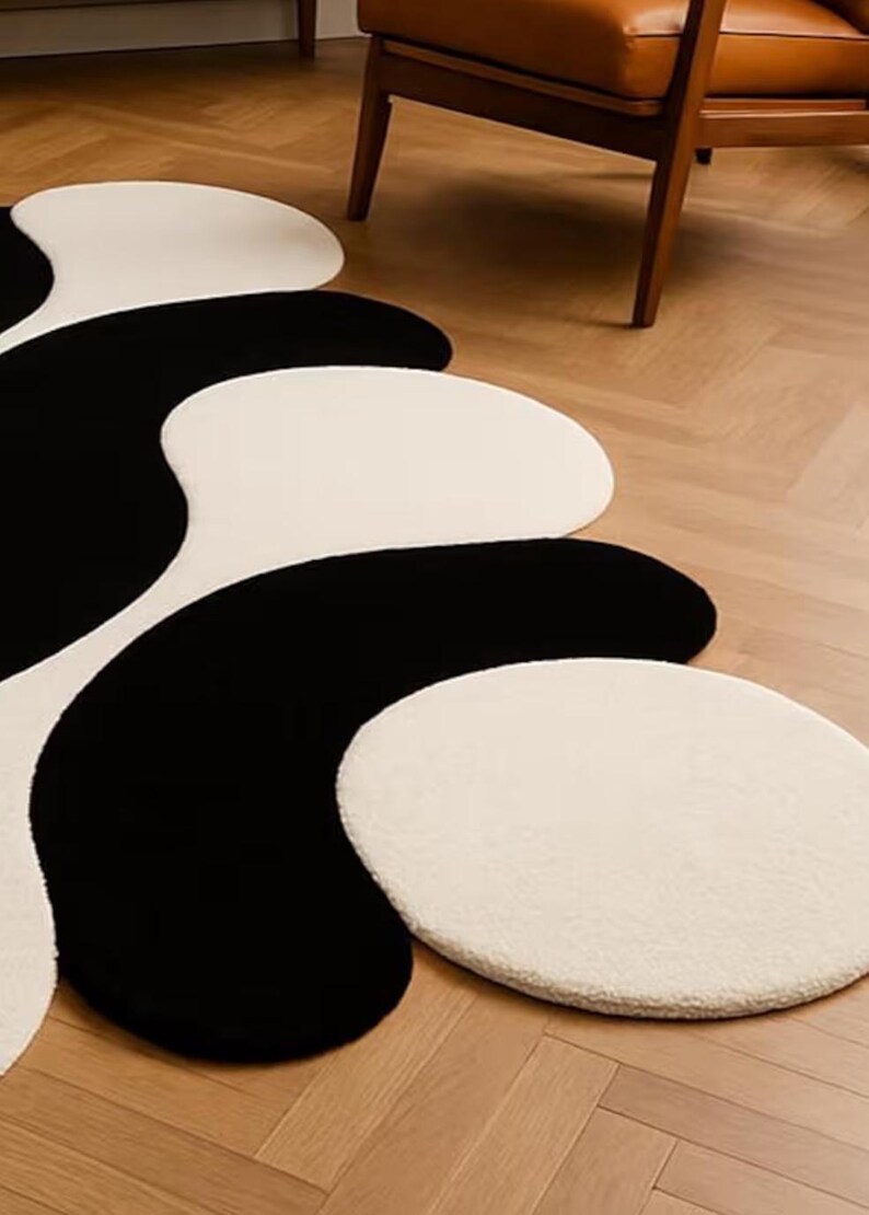 May include: A modern area rug with a unique design. The rug features a flowing pattern of black and off-white shapes. The shapes are soft and organic, creating a visually interesting and stylish floor covering. The rug is placed on a wooden floor.