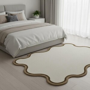 May include: A cream-colored, cloud-shaped rug with a brown border sits on a light wood floor in a bedroom. A bed with a gray headboard, white bedding, and decorative pillows is in the background.