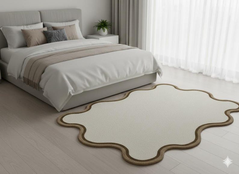 May include: A cream-colored, cloud-shaped rug with a brown border sits on a light wood floor in a bedroom. A bed with a gray headboard, white bedding, and decorative pillows is in the background.