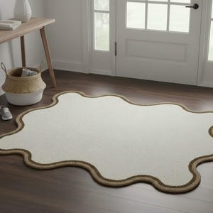 May include: A cream-colored, irregularly shaped rug with a brown border. The rug is placed on a dark wood floor near a doorway. A small wooden table, a basket, and a pair of shoes are also visible.