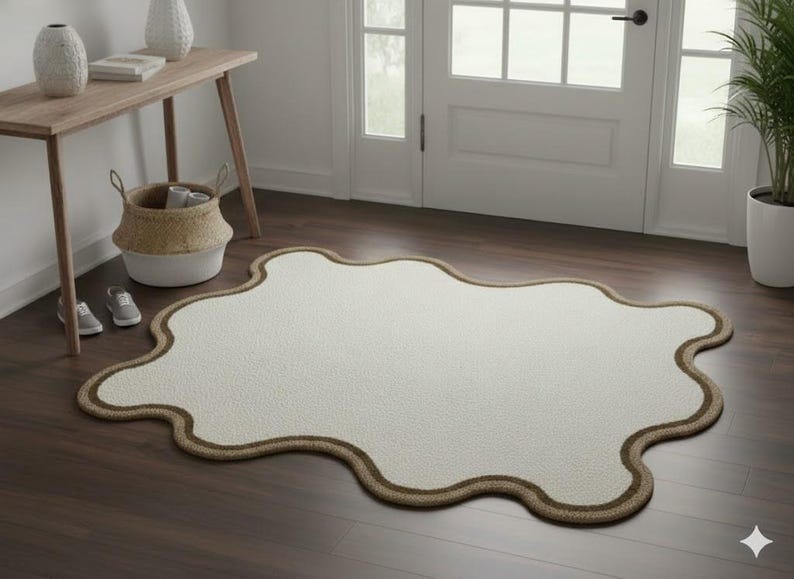 May include: A cream-colored, irregularly shaped rug with a brown border. The rug is placed on a dark wood floor near a doorway. A small wooden table, a basket, and a pair of shoes are also visible.