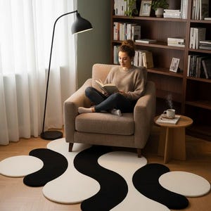 May include: A modern living room scene featuring a black and white abstract rug. A woman is seated in a beige armchair, reading a book. A black floor lamp and a wooden bookshelf complete the cozy interior.