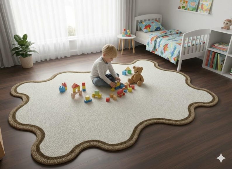 May include: A cream-colored, irregularly shaped rug with a brown border. A young child is playing with wooden blocks and a teddy bear on the rug. A white bed with dinosaur-themed bedding is in the background.