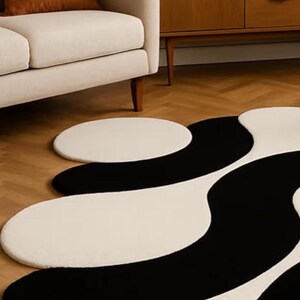 May include: A modern, abstract area rug with a black and white wavy pattern. The rug is on a hardwood floor, and a cream-colored sofa and wooden cabinet are in the background. The rug's design adds a contemporary touch to the room.