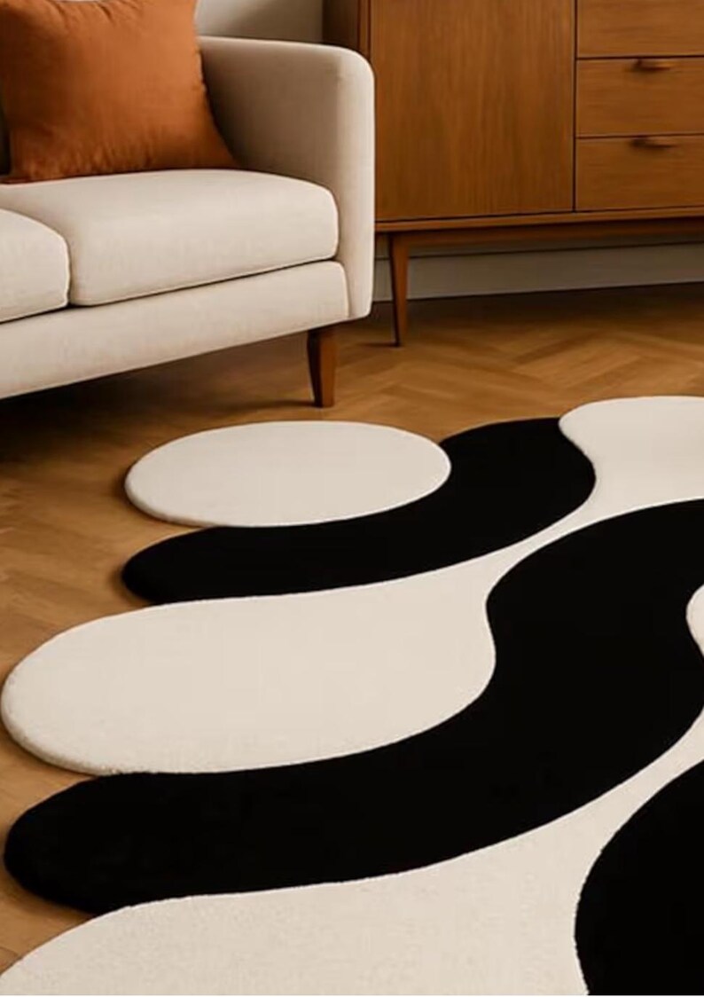 May include: A modern, abstract area rug with a black and white wavy pattern. The rug is on a hardwood floor, and a cream-colored sofa and wooden cabinet are in the background. The rug's design adds a contemporary touch to the room.