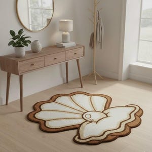 Könnte beinhalten: Ein muschelförmiger Teppich mit Perlendesign. Der Teppich hat ein weißes Innere mit einem braunen Rand und einer Perle in der Mitte. Er liegt auf einem hellen Holzboden und verleiht dem Raum einen Hauch von Küstendekor.