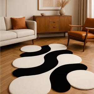 May include: A modern area rug with an abstract design in black and off-white. The rug features flowing, organic shapes and is placed on a hardwood floor. The room also includes a sofa, chairs, and a wooden cabinet, creating a stylish living space.