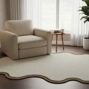 May include: A cream-colored, uniquely shaped rug with a brown border sits on a dark wood floor. A beige armchair and a floor lamp are to the left. A small wooden side table with a mug and books is near a window with white curtains.