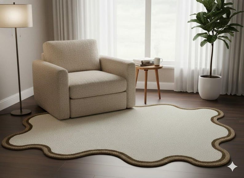 May include: A cream-colored, uniquely shaped rug with a brown border sits on a dark wood floor. A beige armchair and a floor lamp are to the left. A small wooden side table with a mug and books is near a window with white curtains.