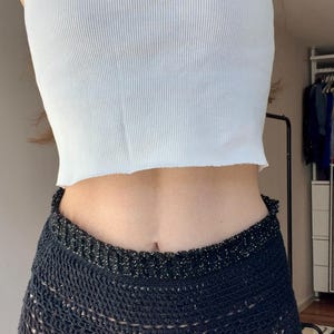 Könnte beinhalten: Ein weißes geripptes Tanktop und schwarze Häkelshorts. Die Shorts haben einen dekorativen, glitzernden Besatz an Bund und Saum. Das Outfit ist lässig und für warmes Wetter geeignet. Die Shorts sind aus Häkelstoff.