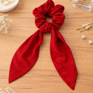 May include: A bright red scrunchie with long, fabric tails. The hair accessory is made of a soft material and is designed to hold hair in place. The scrunchie is a bold fashion statement.