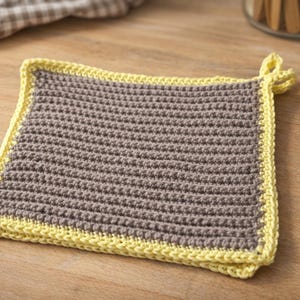 Könnte beinhalten: Quadratischer, handgehäkelter Topflappen in Grau mit gelbem Rand. Der Topflappen hat eine Schlaufe zum Aufhängen und besteht aus strukturiertem Garn. Ein Küchenaccessoire zum Schutz vor Hitze.