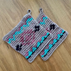 Op de afbeelding: Twee vierkante, gehaakte pannenlappen. De pannenlappen zijn voornamelijk grijs met turquoise, donkerpaarse en roze strepen en accenten. Elke pannenlap heeft een kleine lus om op te hangen.