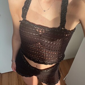 Könnte beinhalten: Ein dunkelbraunes Häkeltop und ein passendes Minirock-Set. Das Neckholder-Top und der Rock haben ein strukturiertes, offen gewebtes Design. Das Set besteht aus einem glänzenden, dunkelbraunen Material, perfekt für ein Sommeroutfit.