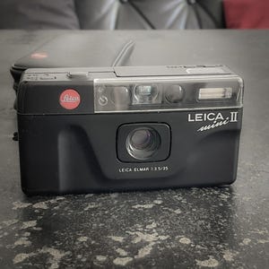 Op de afbeelding: Zwarte Leica Mini II camera met een rood Leica logo. De camera heeft een lens met de tekst "LEICA ELMAR 1:3.5/35" en een flitser. Een zwarte hoes staat op de achtergrond.