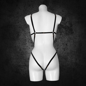May include: Black lingerie body harness with gold-coloured hardware. The harness features multiple straps that connect at the shoulders, back, and waist. The garment is displayed on a white mannequin against a dark grey background.